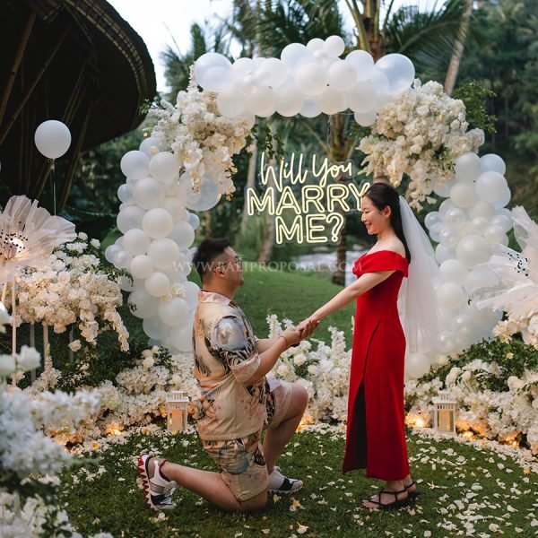 Man proposing to a woman in red under a white balloon arch with a neon Will You Marry Me sign