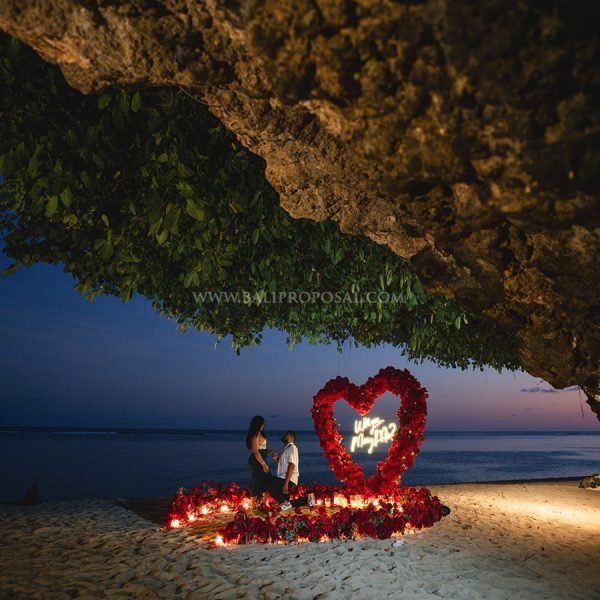 Couple holding hands in a flower arch with a Marry Me sign and rose petals on the beach