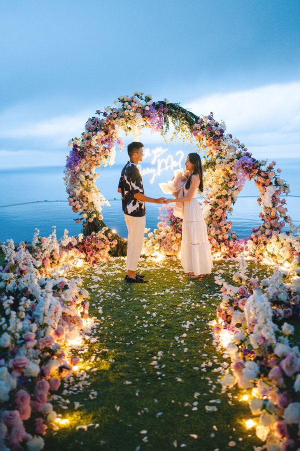Couple holding hands in front of a round flower arch with a neon Will You Marry Me sign by the ocean