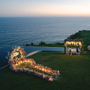 Aerial view of a flower heart and a candlelit dinner table on a grass cliff above the ocean