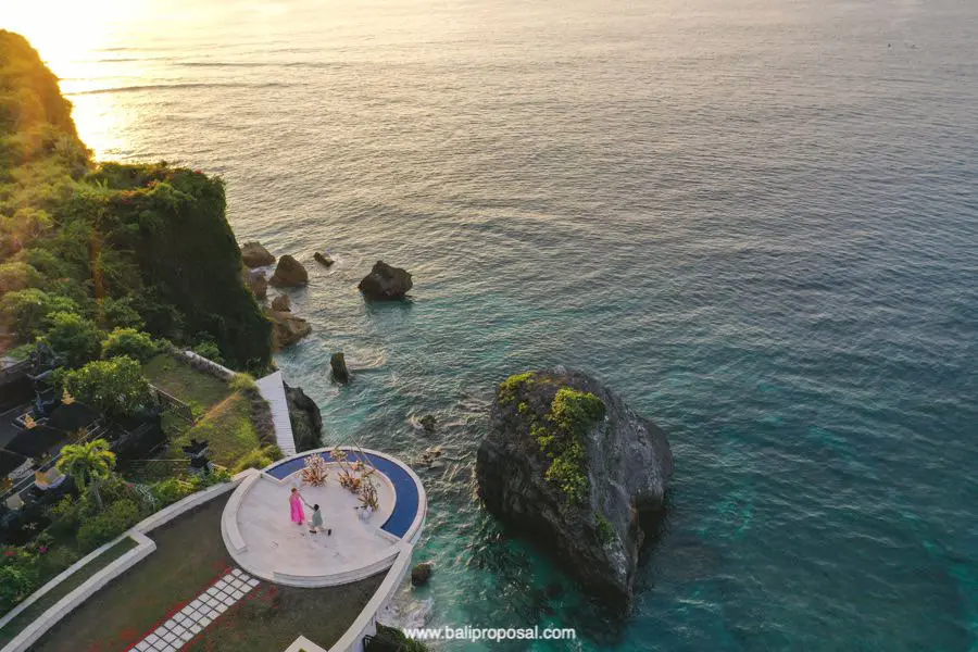 Aerial view of David and Paris on a round cliffside platform above the ocean at sunset